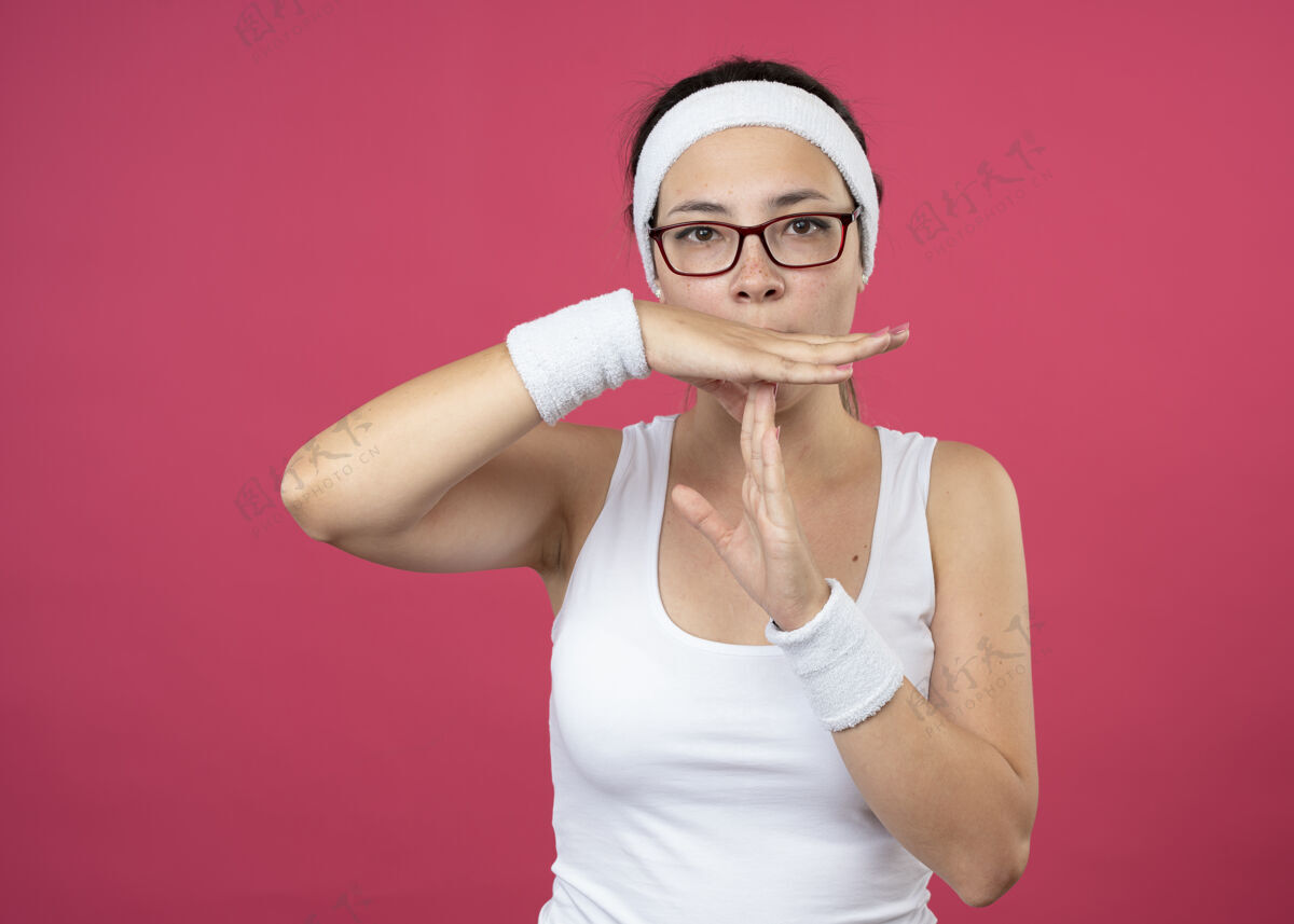 佩戴自信的年轻运动女性戴着眼镜戴着头带和腕带手势暂停标志隔离在粉红色的墙上人手势年轻