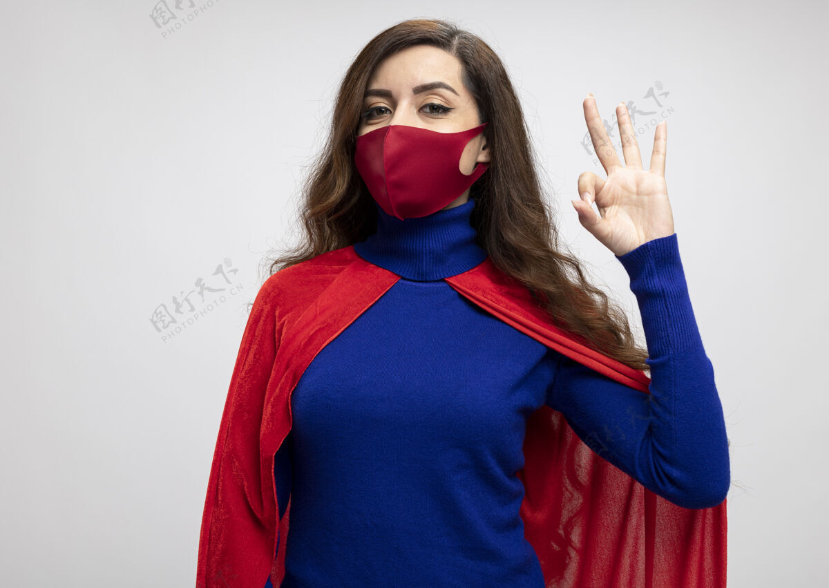 手高兴的女超人穿着红色斗篷戴着红色防护面具手势ok手势隔离在白色墙上保护脸人