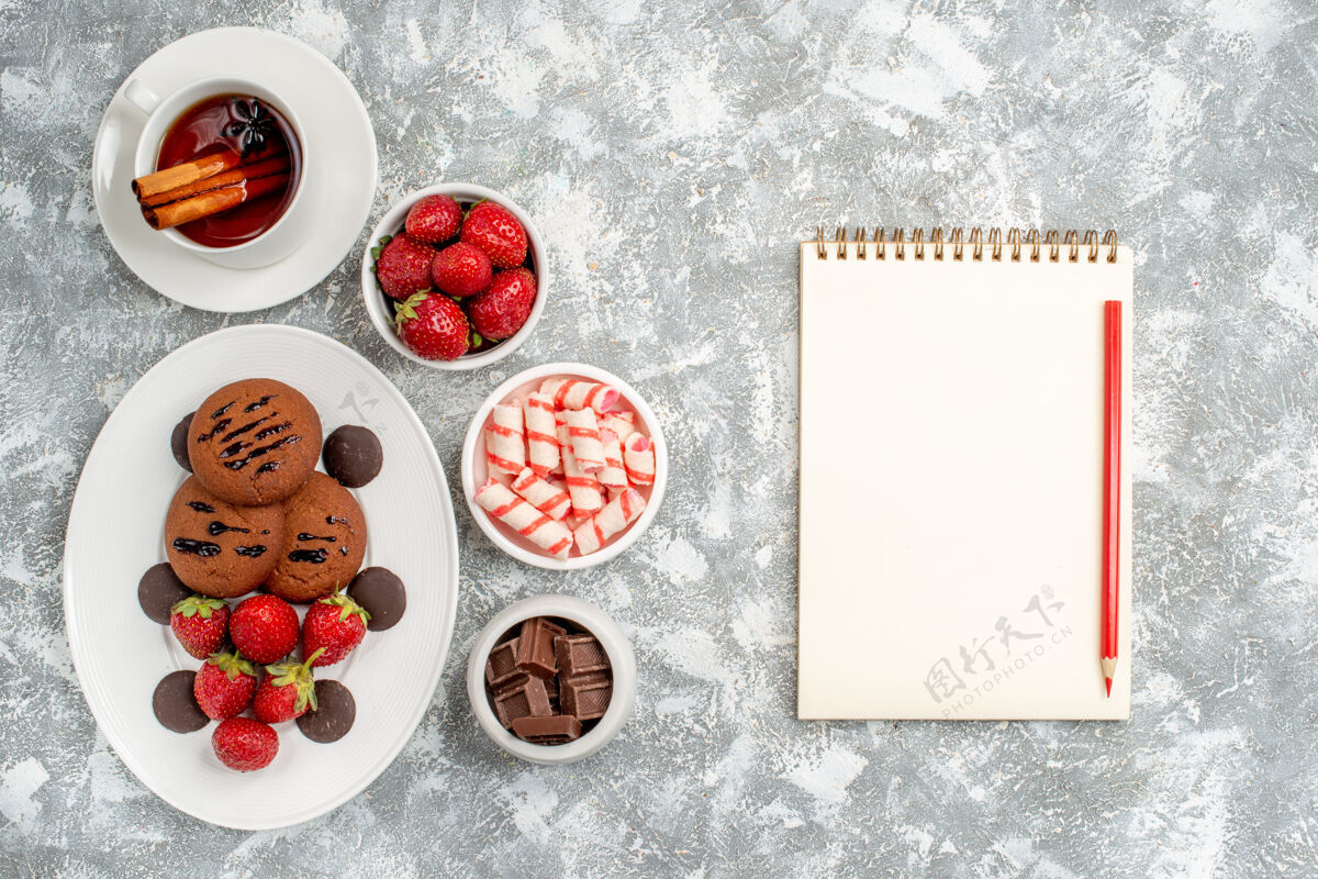 浆果顶视饼干草莓和圆形巧克力放在椭圆形的盘子里 周围是糖果 草莓 巧克力 肉桂茶和笔记本铅笔放在灰白色的桌子上食物草莓铅笔
