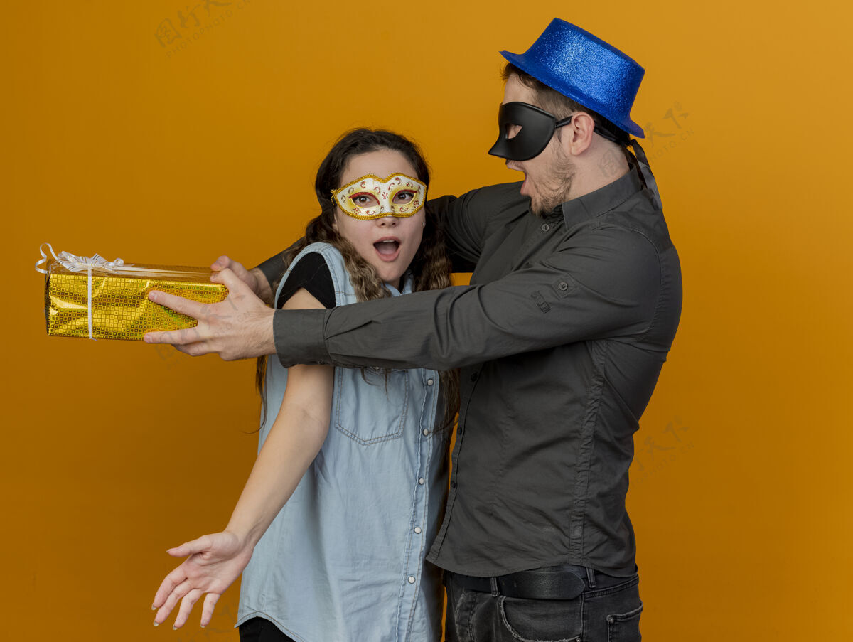男人快乐的年轻派对情侣戴着化装舞会eue面具的家伙拿着礼品盒在女孩身后孤立在橙色化装舞会礼物情侣
