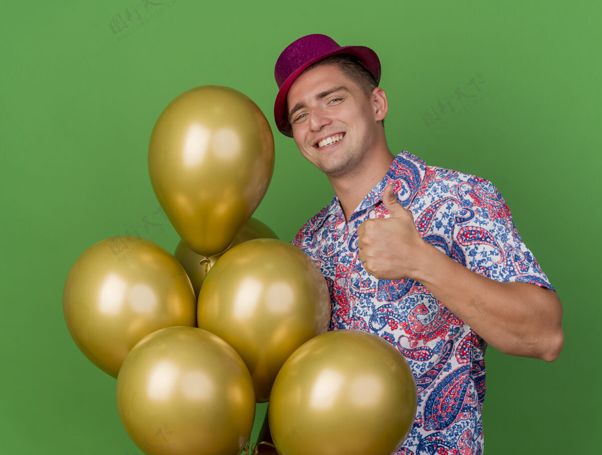 派对微笑着的年轻人戴着粉红色的帽子 拿着气球 在绿色的草地上孤立地竖起大拇指男人年轻展示