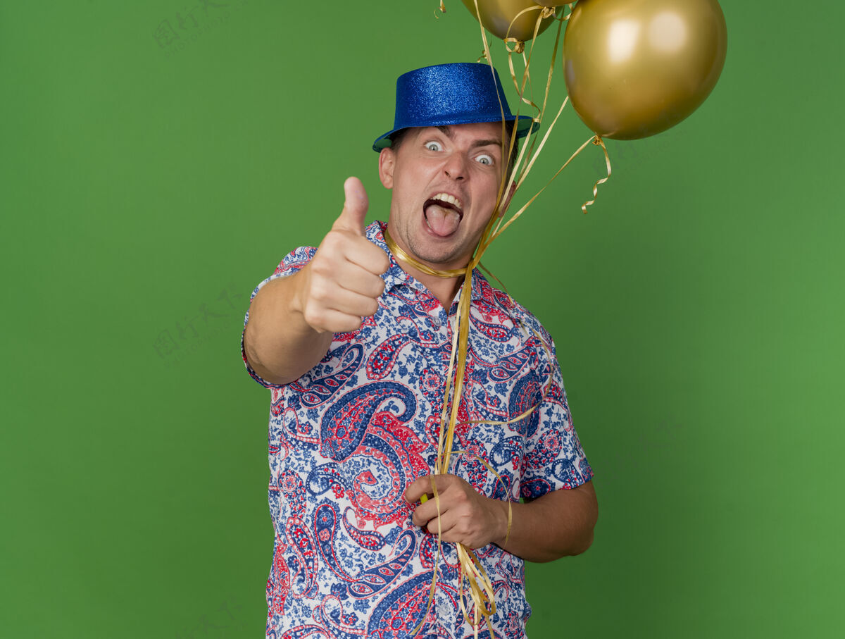 气球快乐的年轻人戴着蓝色帽子 脖子上绑着气球 拇指朝上 舌头孤立在绿色上年轻人抱着绿色