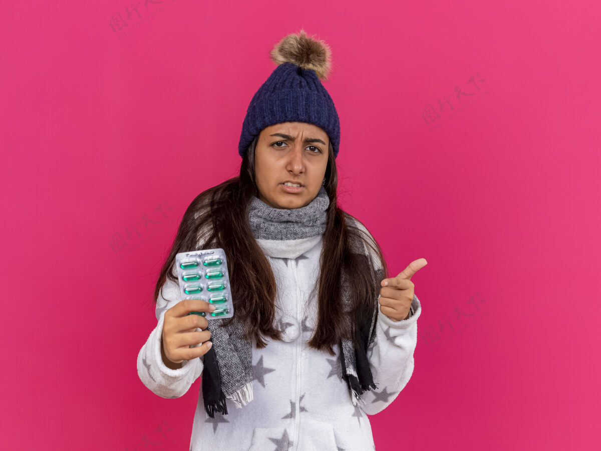 手势戴着冬天的帽子 戴着围巾 拿着药片 显示你在粉红色上孤立的姿势的未被释放的生病的年轻女孩帽子生病药片