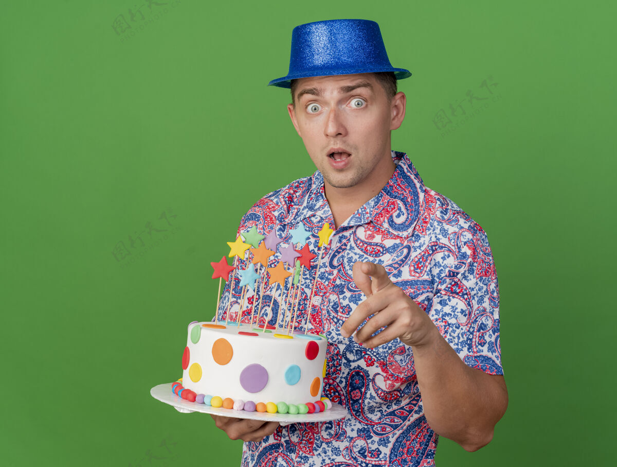 派对惊讶的年轻人戴着蓝色的帽子拿着蛋糕在绿色上展示你的姿势蓝色男人举行