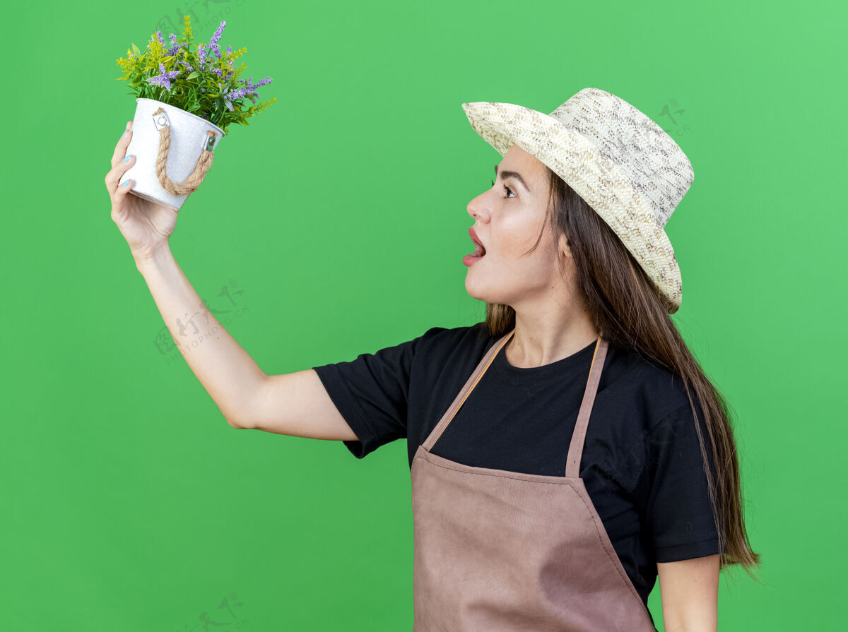 提高站在侧视图上美丽的园丁身着制服的女孩戴着园艺帽抬头望着隔离在绿色花盆里的花朵景观绿色穿