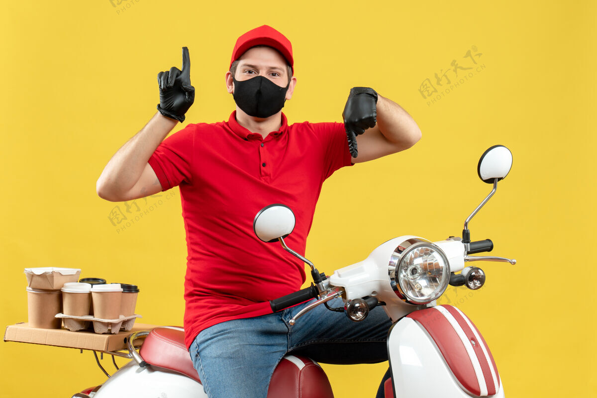 年轻人俯视图中的年轻人穿着红色上衣 戴着帽子手套 戴着医用面罩 坐在踏板车上 在黄色背景上上上下竖起大拇指黄色滑板车交付