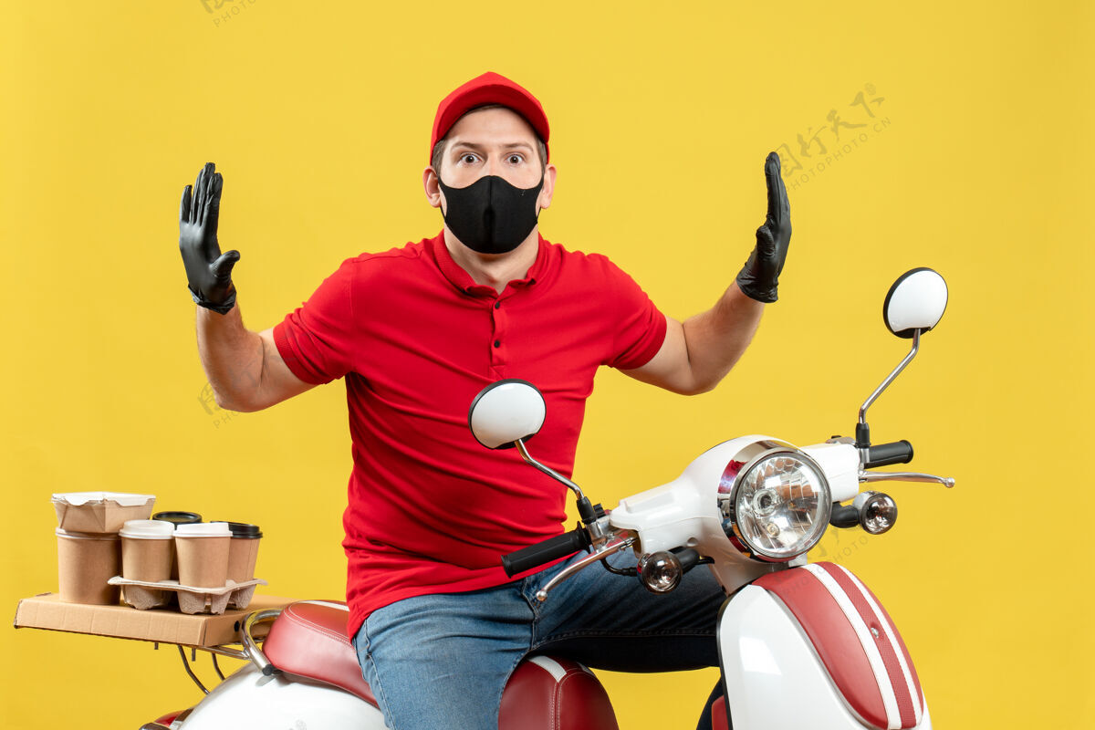 背景俯视图：戴着医用口罩 身穿红色上衣 戴着帽子手套的年轻人坐在踏板车上 在黄色背景下感到震惊人摩托车医学