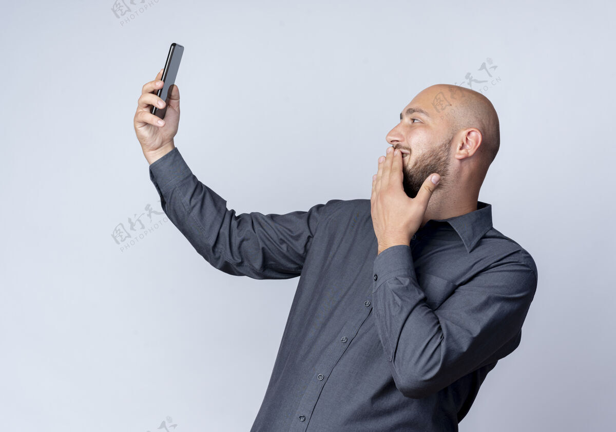 中心微笑着的年轻秃头呼叫中心的男子拿着手机看着把手放在嘴唇上隔离在白色的墙上白脸公民