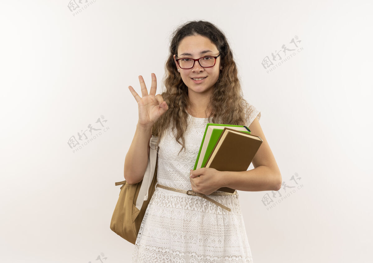 背高兴的年轻漂亮的女学生戴着眼镜 背着书包拿着书做ok的标志隔离在白色的墙上市民眼镜白
