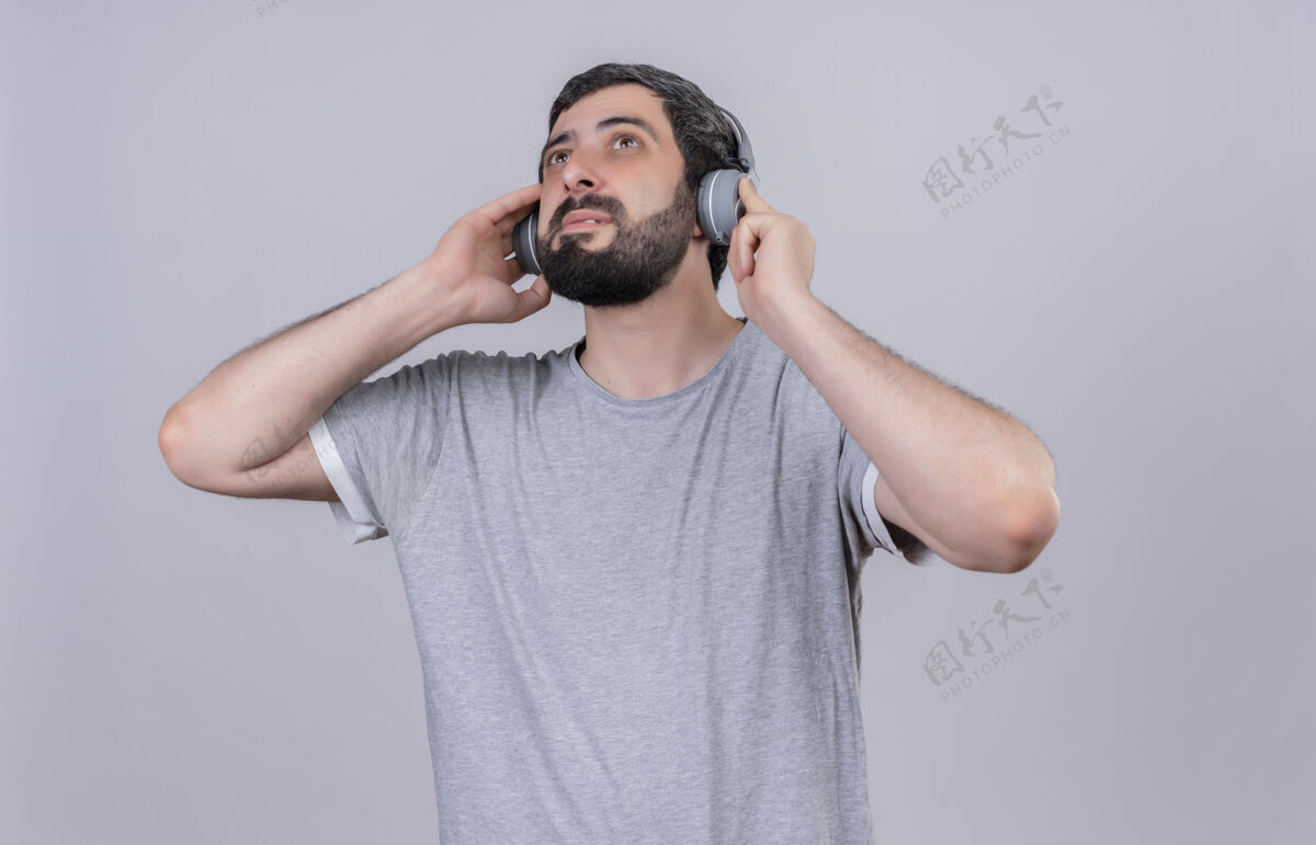 脸高兴的年轻帅哥戴着耳机听音乐 抬起头用手把耳机隔离在白墙上请穿着姿势