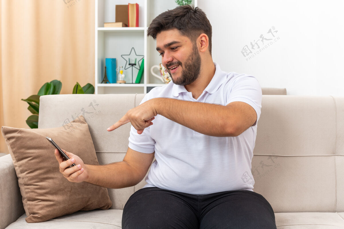 举行穿着休闲服的年轻人拿着智能手机 用食指指着它 快乐而积极地坐在客厅里的沙发上手机坐着手指