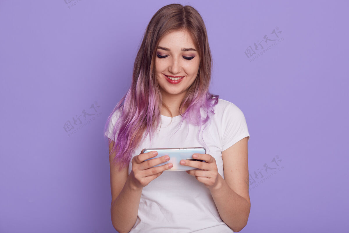 女孩微笑的女性穿着白色t恤 手里拿着智能手机 玩电镀游戏 带着可爱的微笑看着显示器 用手机 在紫色的墙上摆出孤立的姿势女人打字人