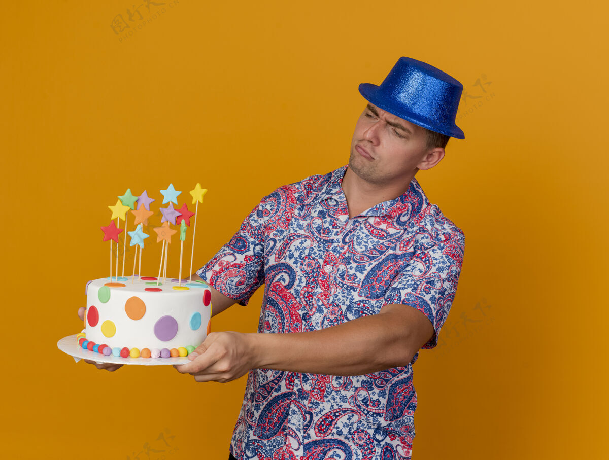 派对迷茫的年轻人戴着蓝色的帽子 拿着蛋糕站在橙色的一边小伙子侧板蛋糕