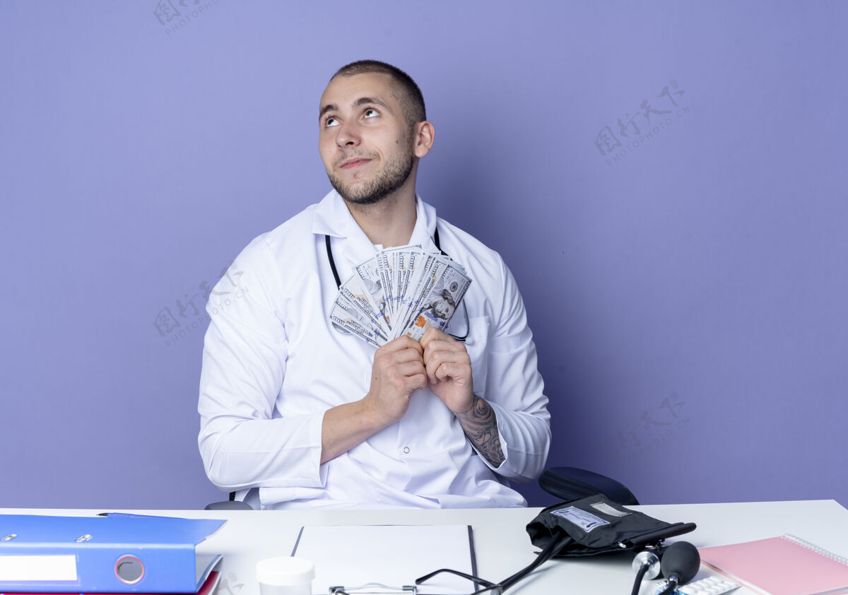 人年轻的男医生穿着医用长袍和听诊器坐在办公桌旁 手里拿着工作工具 手里拿着钱 孤零零地仰望着紫色的墙上男工作紫色