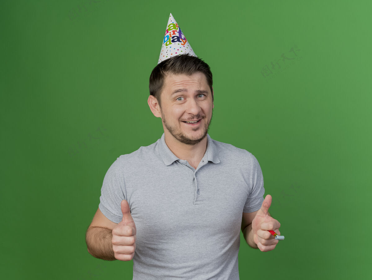 男人微笑的年轻人戴着生日帽 手持哨子 竖起大拇指 孤零零地站在草地上穿派对表演