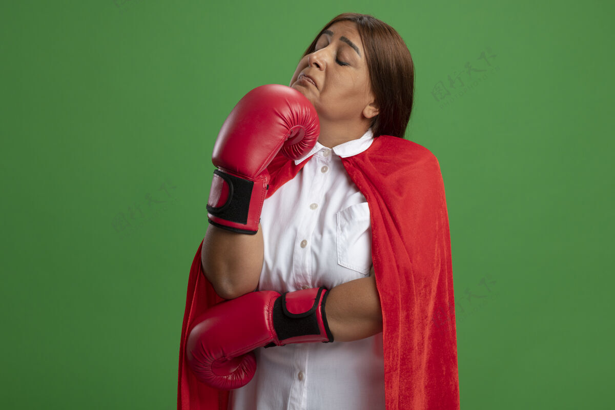 女性体贴的中年女超级英雄闭着眼睛戴着拳击手套把手放在下巴下隔离在绿色的下巴拳击眼睛