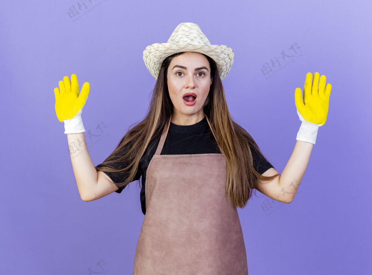 制服惊讶美丽的园丁女孩穿着制服戴着园艺帽子和手套摊开双手孤立在蓝色蔓延园艺帽子