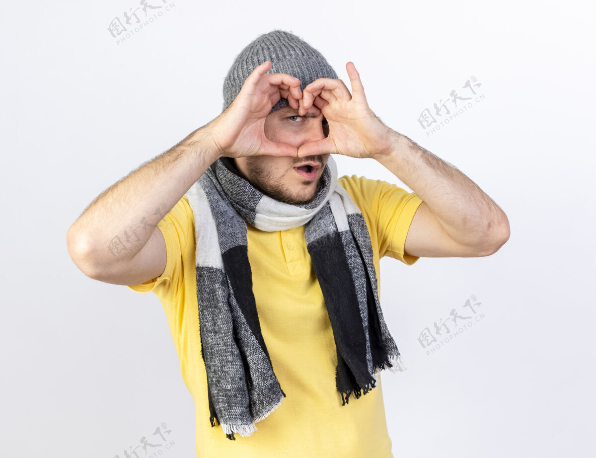 手势自信的年轻金发病夫戴着冬天的帽子和围巾的手势 透过隔离在白色墙上的心形标志看着前方通过脸金发