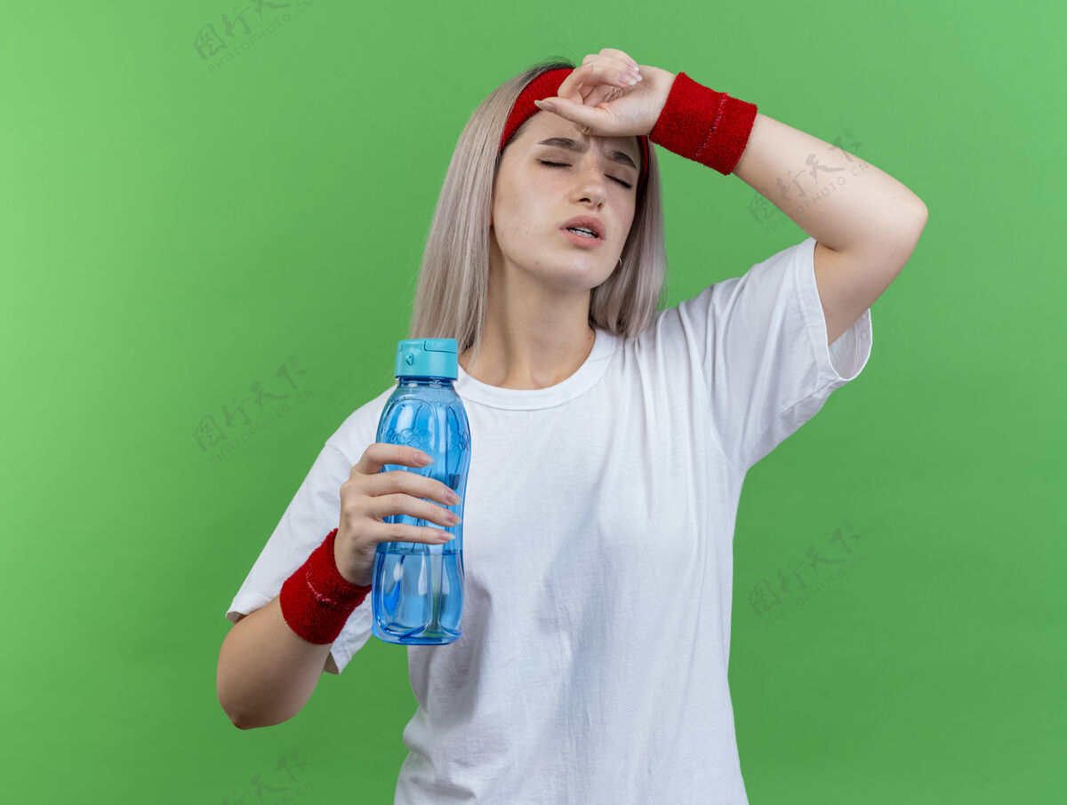 人带着护腕戴着头带和腕带的疼痛的年轻运动女性把手放在额头上 把水瓶隔离在绿色的墙上戴头带抱着