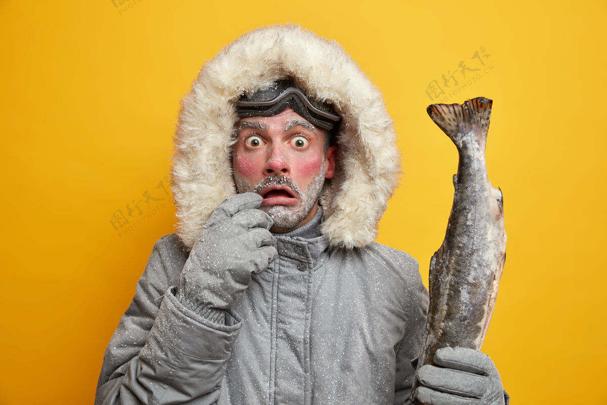 穿户外冬季活动和爱好概念目瞪口呆的男子红着冻僵的脸盯着窃听的眼睛抱着钓到的大鱼穿着暖和的衣服已经成功捕鱼了季节角度兜帽