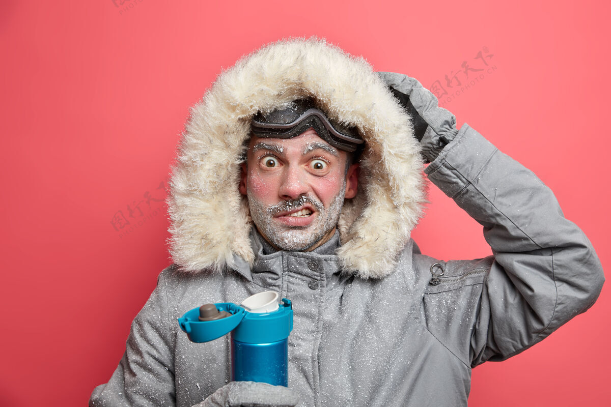 人迷惑不解的男远足者冻僵了脸 在探险中被寒冷的环境所震惊 穿着暖和的夹克 戴着滑雪镜 喝着热饮料夹克封面护目镜