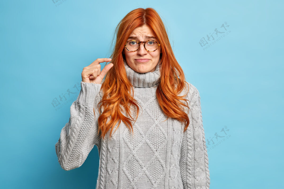 套头衫不高兴的红发年轻欧洲妇女穿着灰色冬季套头衫显示少量的手势显示与手指小尺寸不满意红发姜