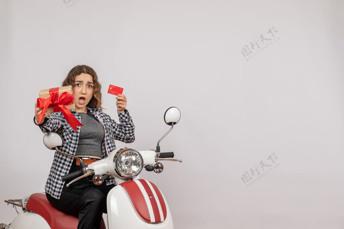 坐着前视图困惑的年轻女子骑着轻便摩托车拿着卡片和礼物在灰色的墙上摩托车人轻便摩托车