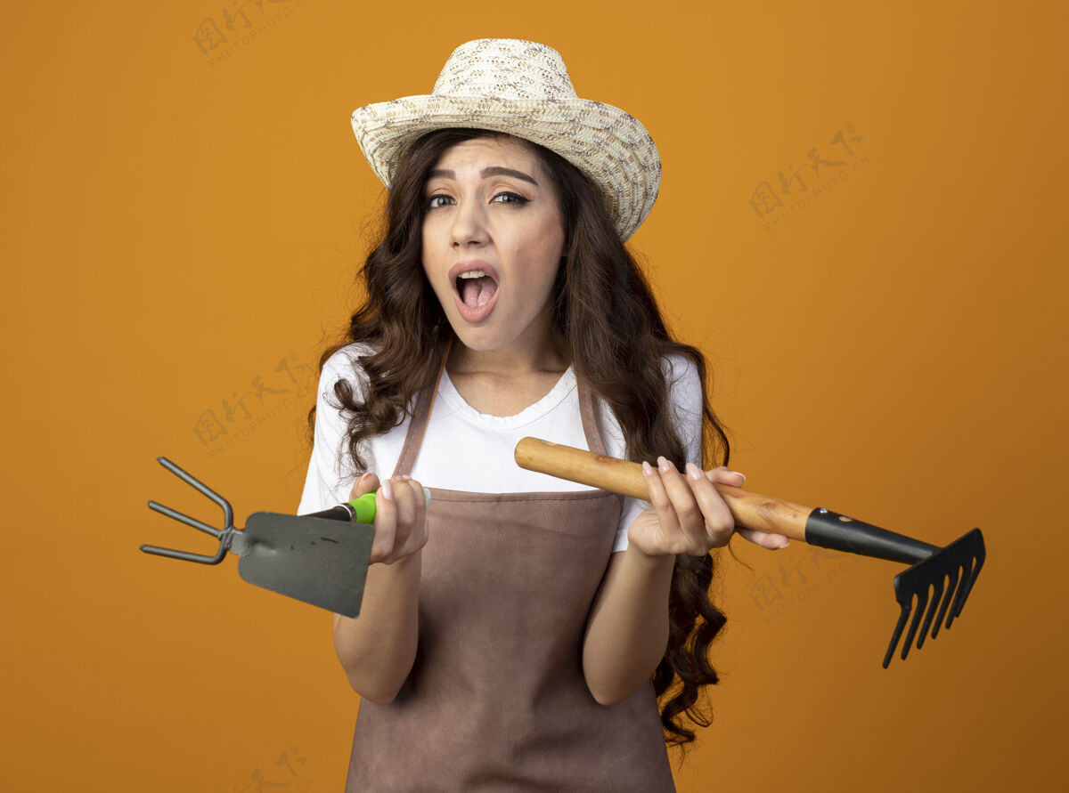 锄头恼怒的年轻女园丁穿着制服 戴着园艺帽 把耙子和锄头耙子隔离在橙色的墙上 留着复制空间园艺帽子花园
