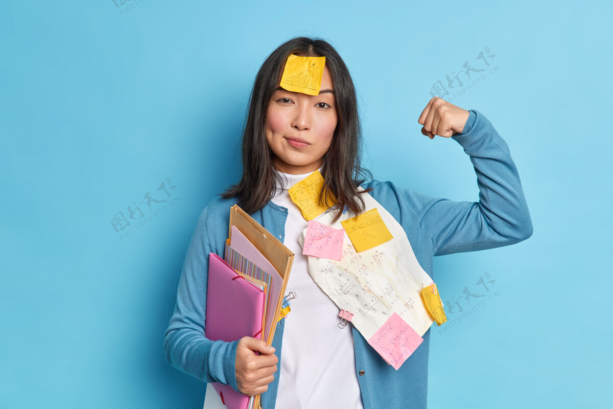 筋疲力尽强大的女学生举起手臂和显示肌肉后 对文凭纸工作感到自信额头上戴着标签举行文件夹女性文件二头肌