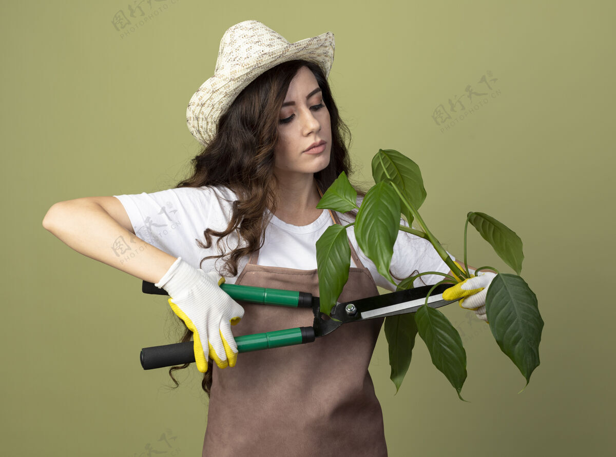 年轻自信的年轻女园丁穿着制服 戴着园艺帽和手套 假装用园丁剪子在橄榄绿的墙上切割植物 并留有复制空间切割园艺假装