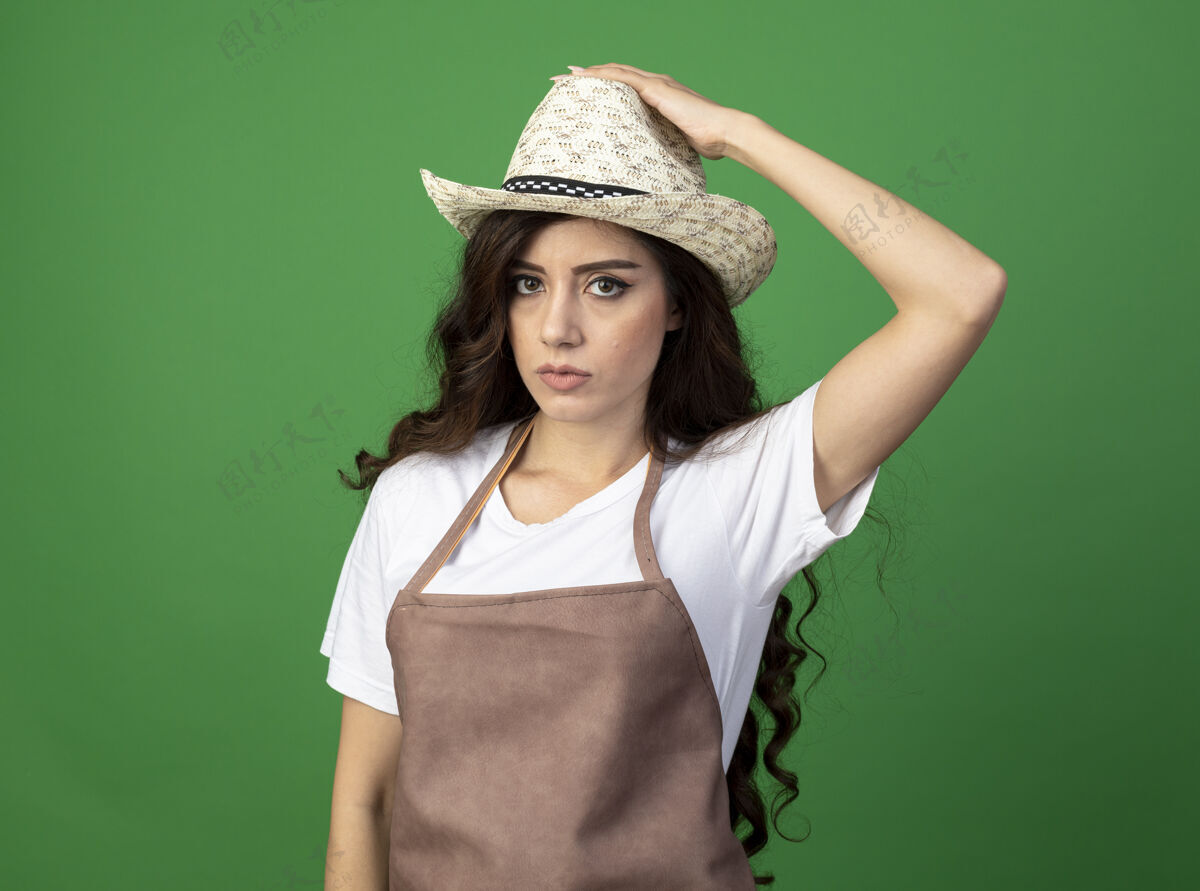 帽子自信的年轻女园丁穿着制服 手戴园艺帽 隔离在绿色墙壁上 留有复制空间花园年轻制服