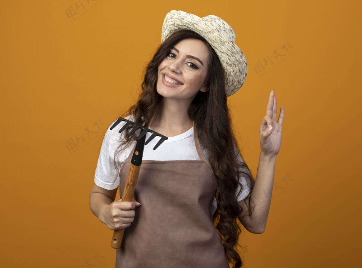 好的身着制服的年轻女园丁面带微笑 戴着园艺帽 拿着耙子 在橘黄色的墙上做着手势“ok”的手势 并留有复制空间帽子花园年轻的