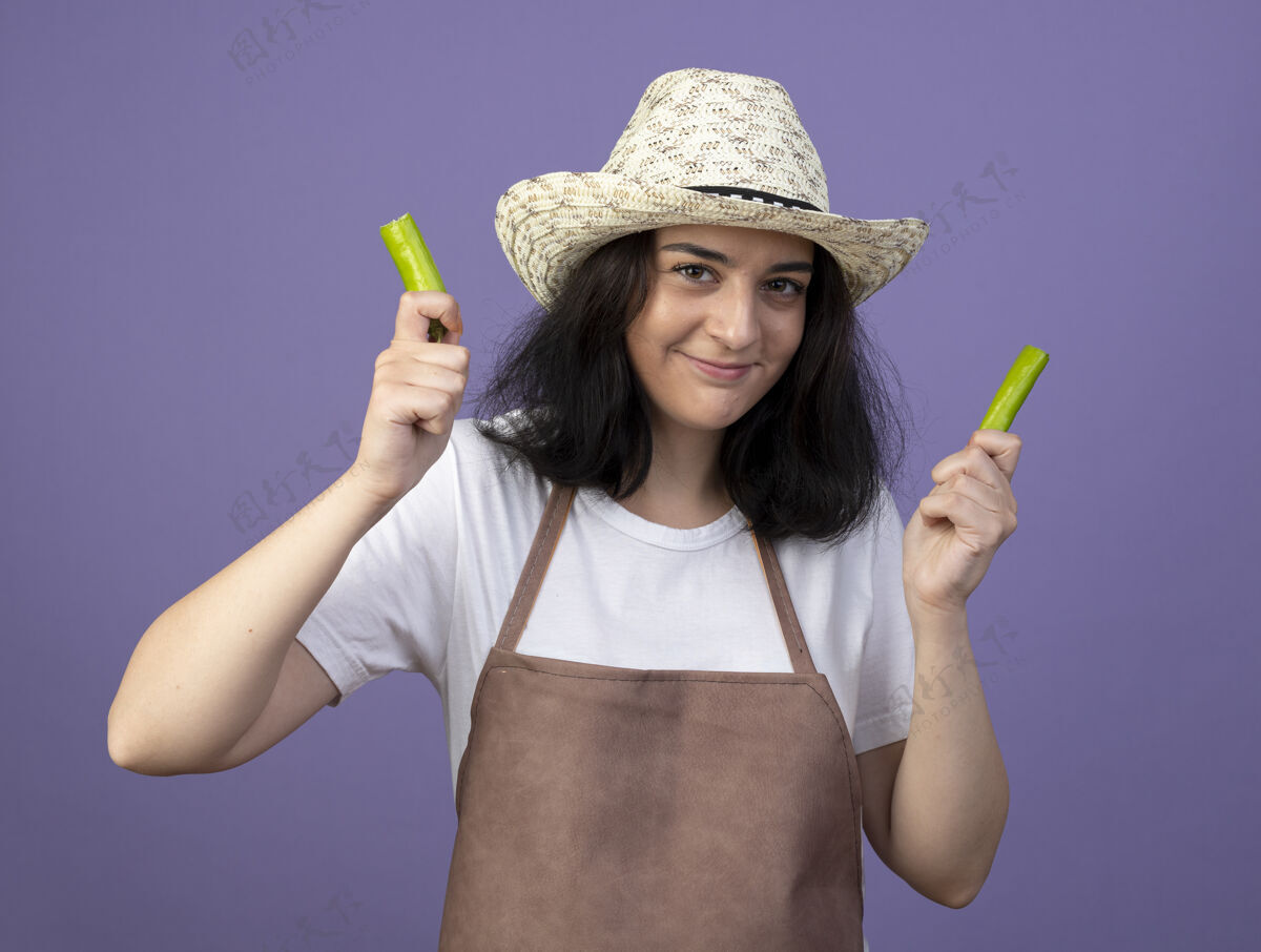 年轻的年轻的黑发女园丁穿着制服 戴着园艺帽 手里拿着一半的辣椒 隔离在紫色的墙上 留着复制空间园艺热的制服