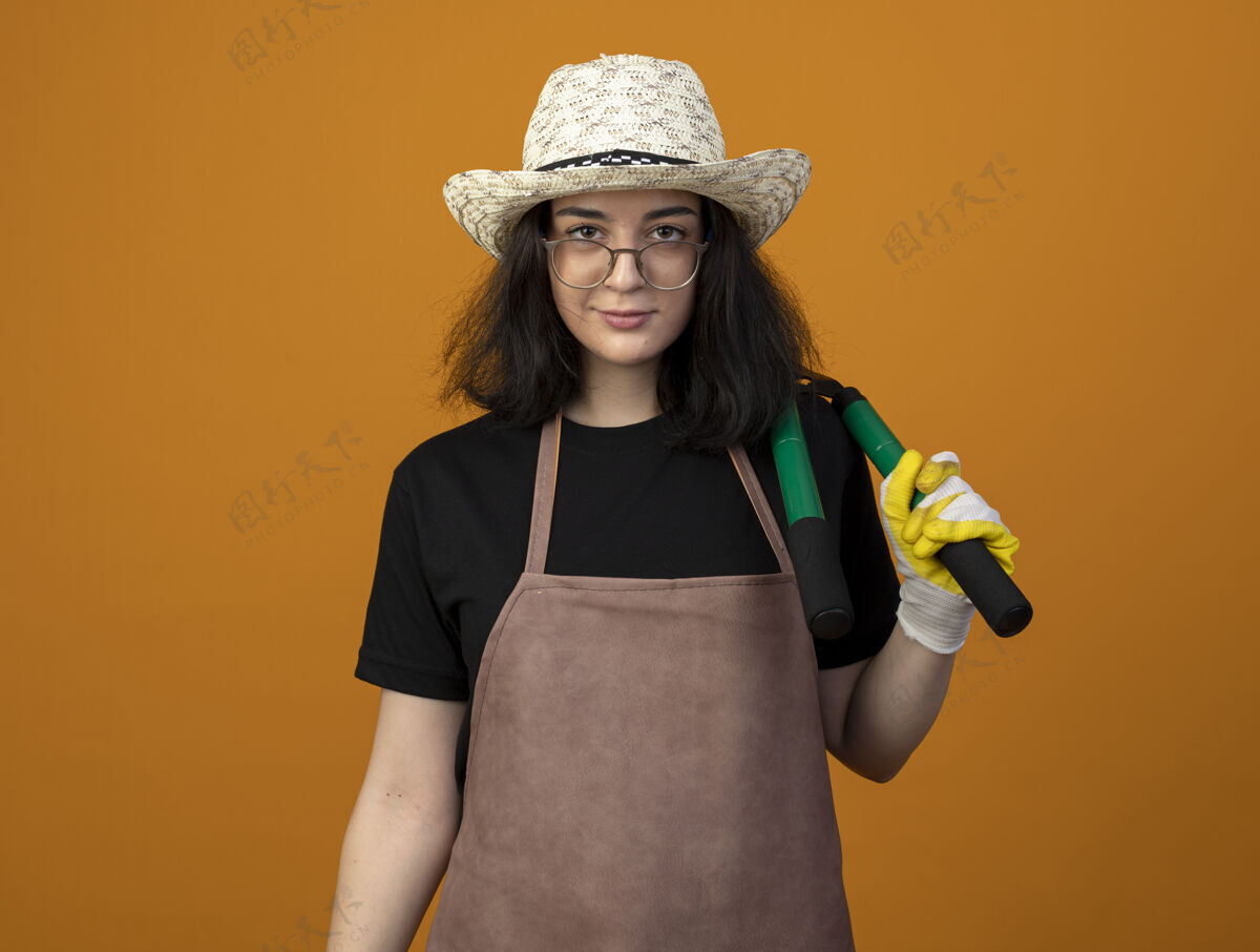 制服自信的年轻黑发女园丁戴着眼镜 穿着制服 戴着园艺帽和手套 肩上拿着园艺剪 隔离在橙色的墙上 留有复制空间肩膀园艺年轻人