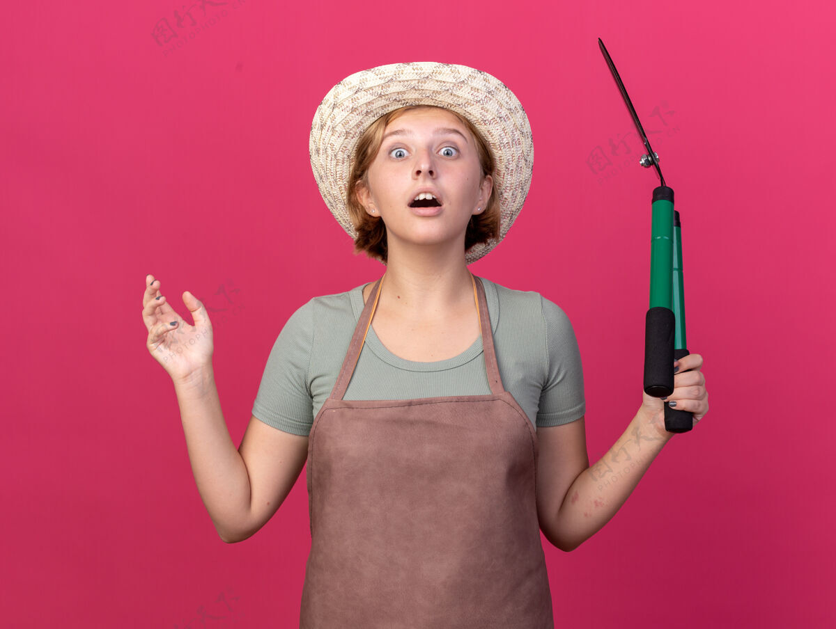 手震惊年轻的斯拉夫女园丁戴着园艺帽子站着 手举园艺剪刀在粉红色剪刀站花园