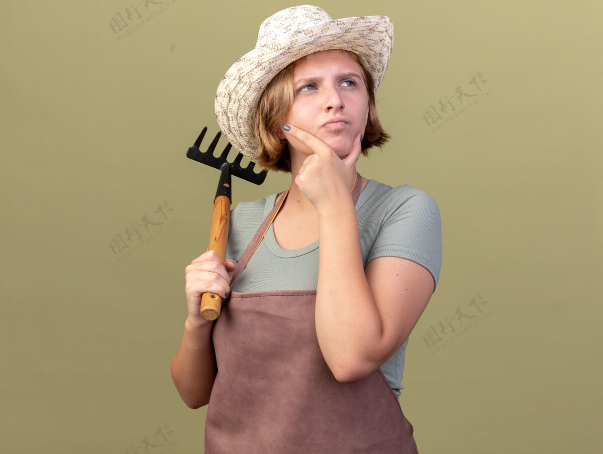 花园体贴的年轻斯拉夫女园丁戴着园艺帽 把手放在下巴上 拿着耙子看着橄榄绿的一面下巴年轻斯拉夫