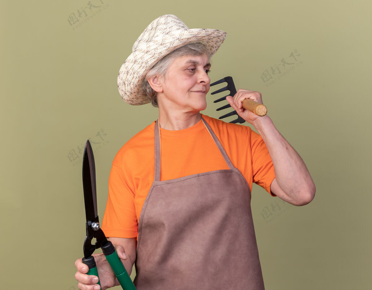花园高兴的上了年纪的女园丁戴着园艺帽手持园艺剪刀和耙子望着橄榄绿的一面剪刀耙子壁板