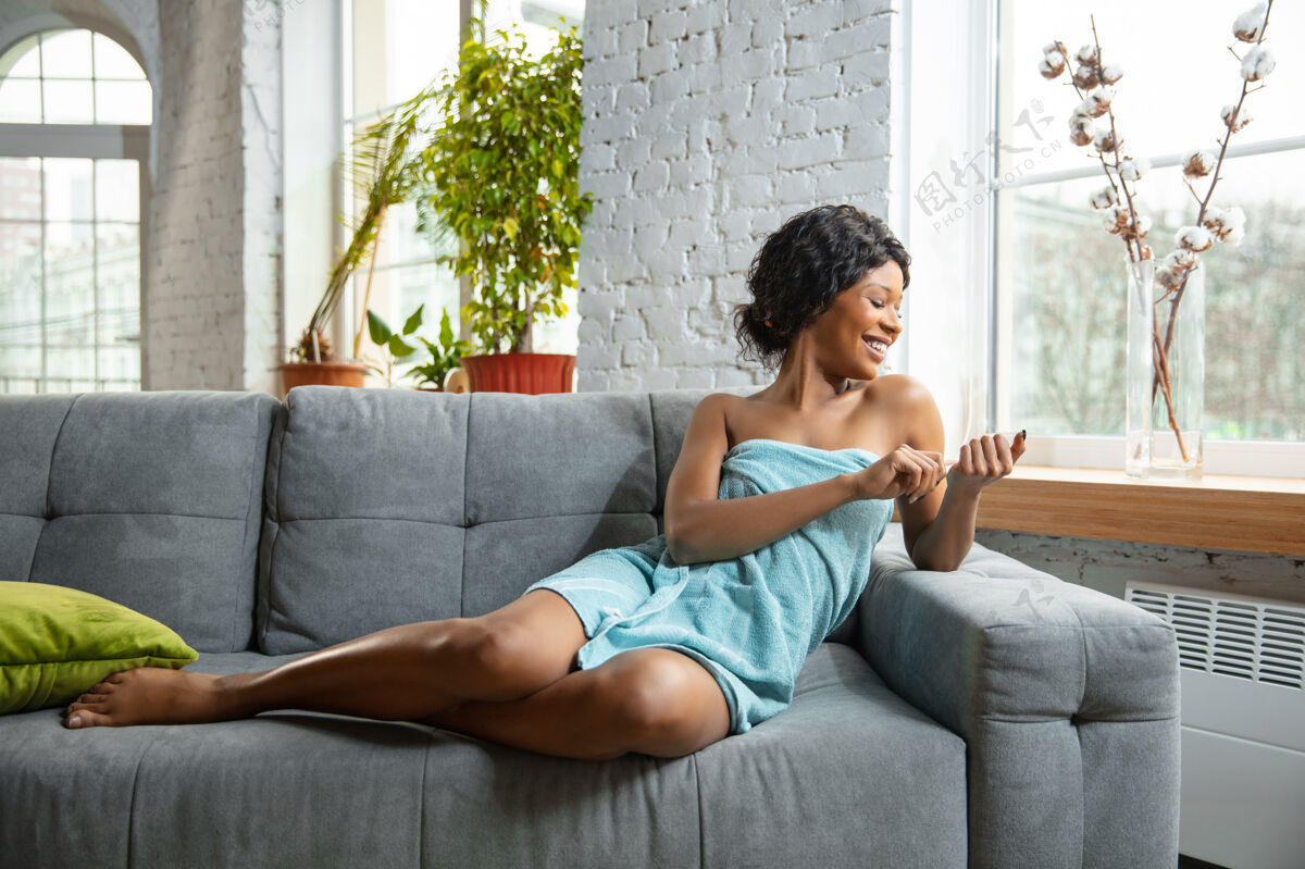 爱美妆日非裔美国妇女用毛巾准备在家做日常护肤坐在沙发上 修指甲 微笑美的概念 自我护理 化妆品 年轻 健康房间脸面部