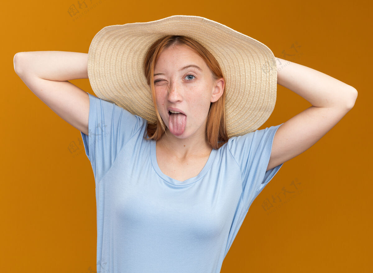 帽子快乐的年轻红发姜女 满脸雀斑 头戴沙滩帽 伸出舌头 把手放在橘子树后面的头上头手喜悦
