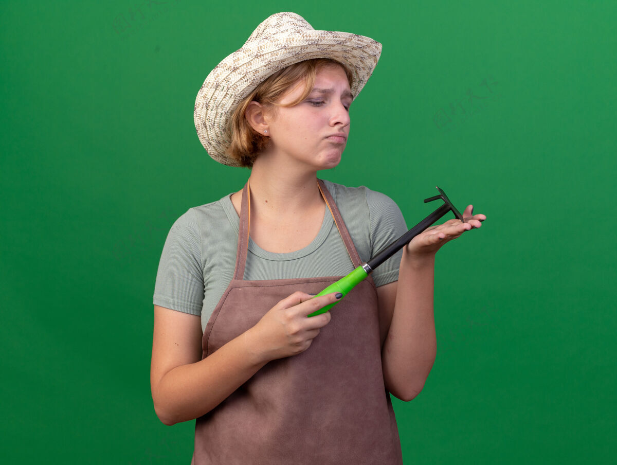 斯拉夫悲伤的年轻的斯拉夫女园丁戴着园艺帽 拿着锄头看耙在草地上年轻园艺悲伤