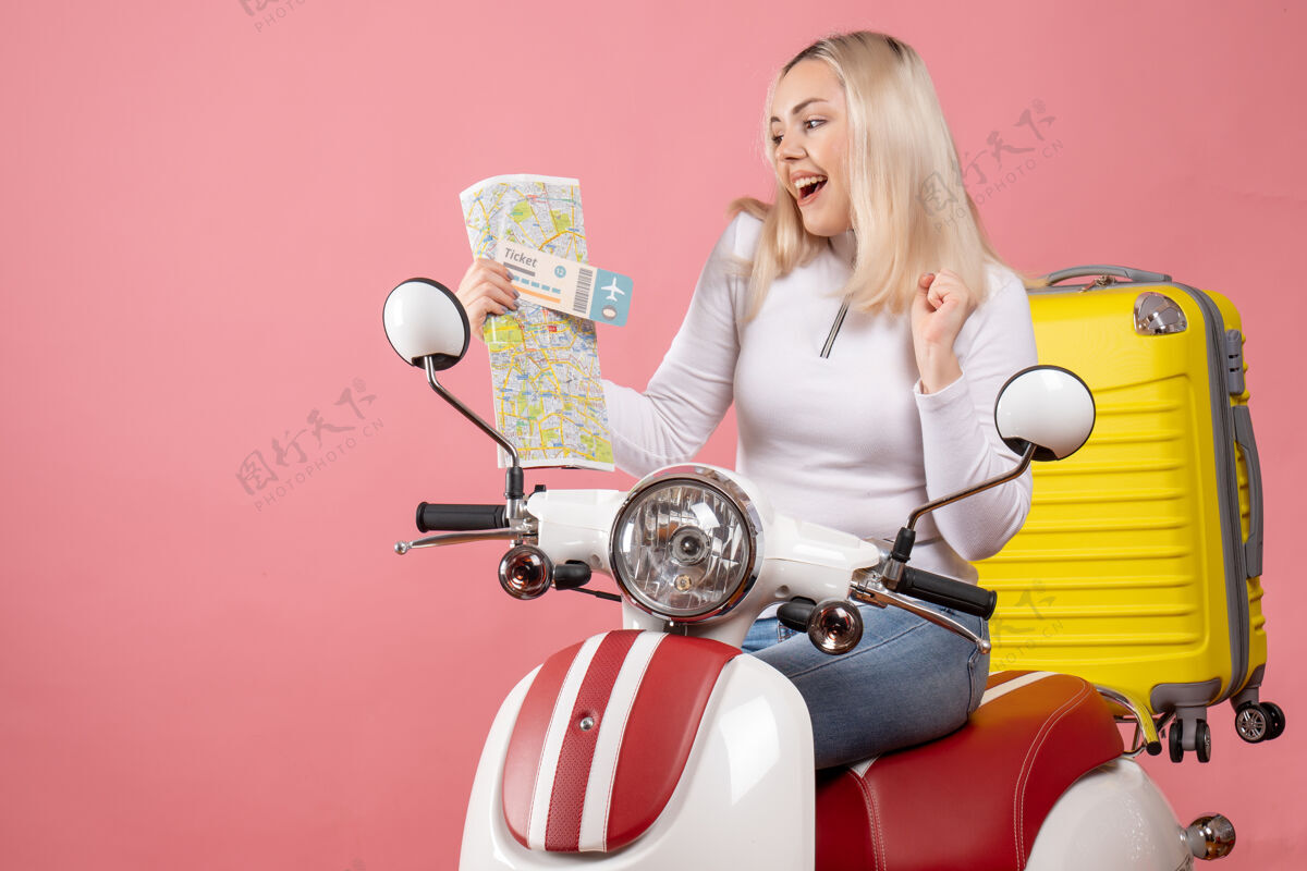 漂亮前视图快乐的金发女孩骑着轻便摩托车拿着票和地图轻便摩托车坐着前面