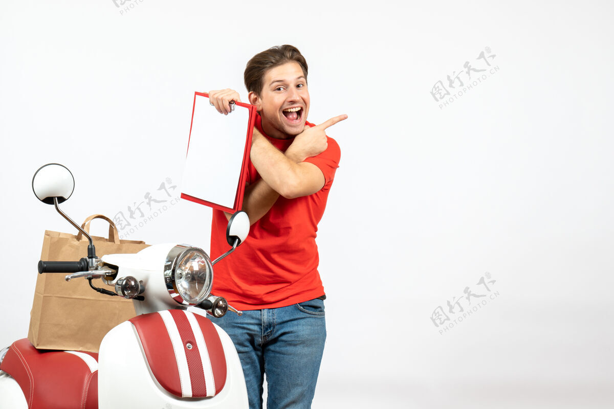 人身着红色制服 微笑着的年轻送货员站在滑板车旁 出示指向白墙上背面的文件送货员年轻人滑板车