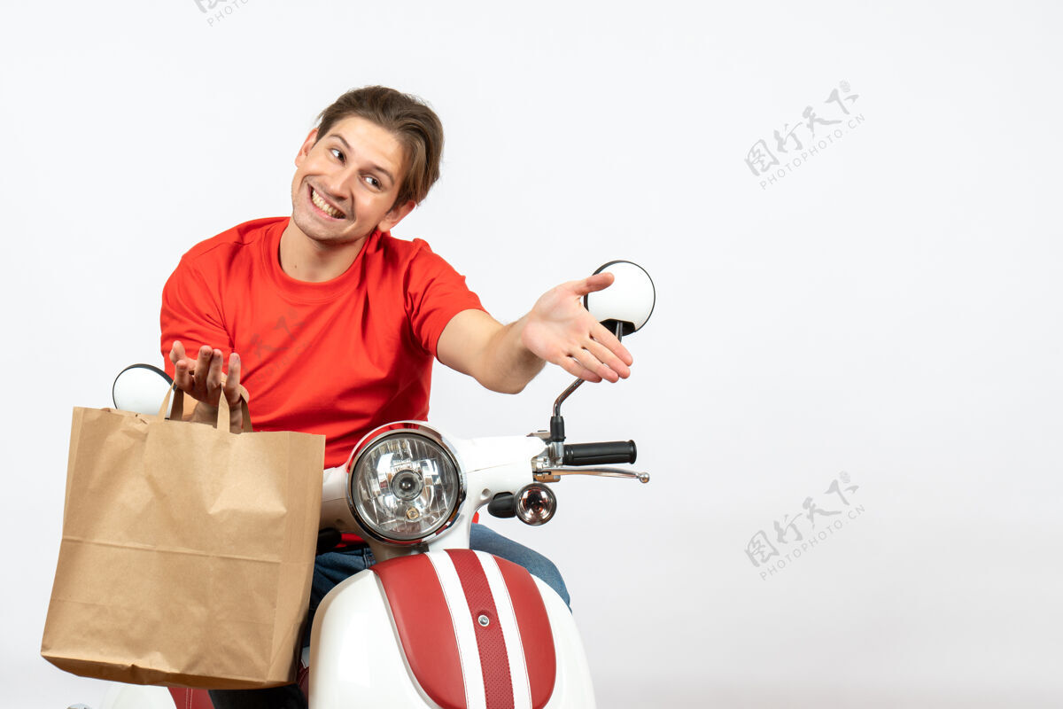 成人身穿红色制服 面带微笑的年轻送货员坐在踏板车上 手里拿着纸袋 在白墙上欢迎大家欢迎人运动
