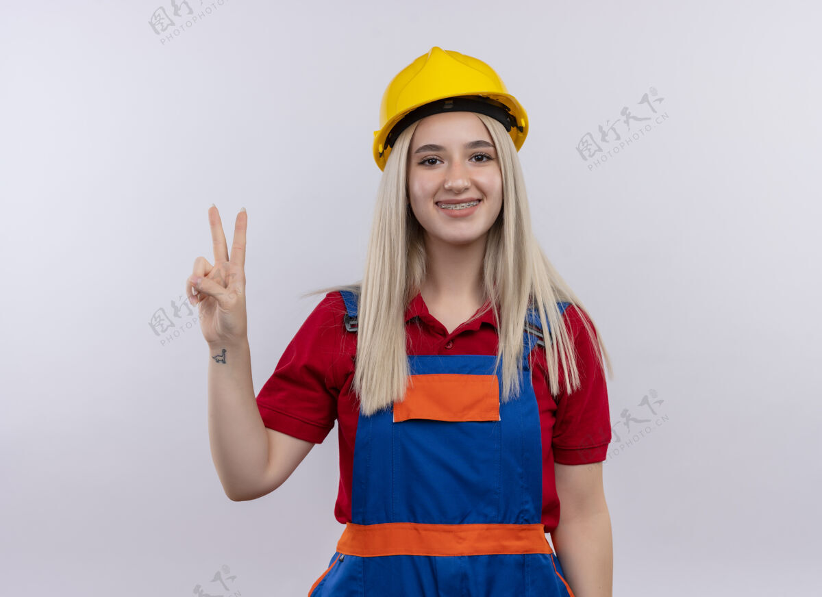 年轻微笑着的年轻金发工程师建筑工女孩穿着制服戴着牙套在与世隔绝的空白处做着和平的手势标志金发牙医