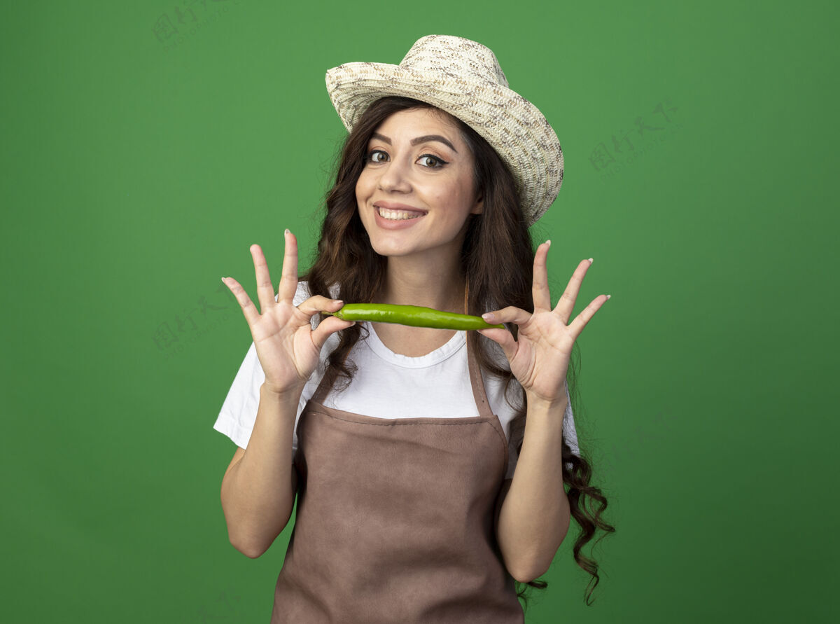 园艺微笑着的年轻女园丁穿着制服 戴着园艺帽 把辣椒隔离在绿色的墙上 留着复制空间年轻的热的胡椒