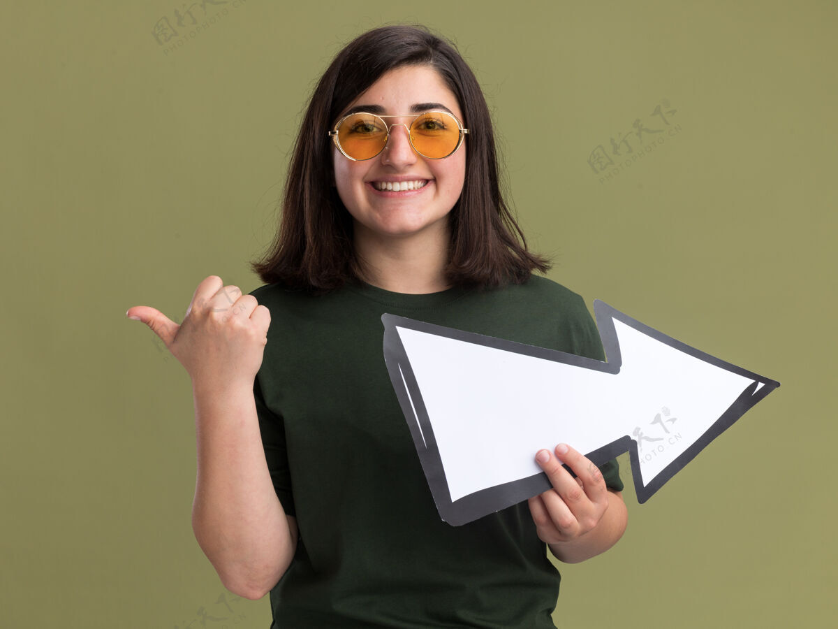 微笑微笑着的年轻漂亮的高加索女孩 戴着太阳眼镜 手持箭头 指着橄榄绿的侧面漂亮指向标志