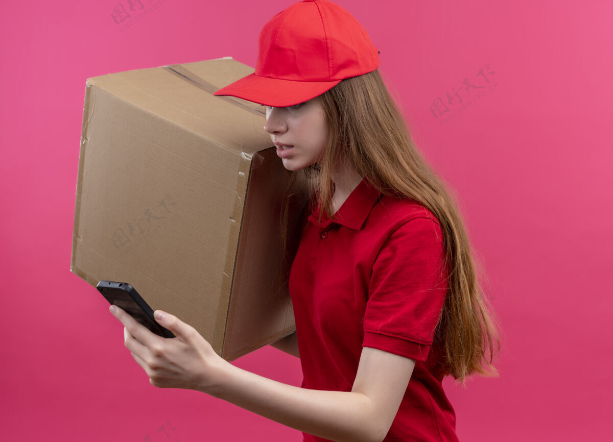 电话印象深刻的年轻送货女孩在红色制服举行的盒子和手机看着手机站在侧面图上孤立的粉红色空间印象送货盒子