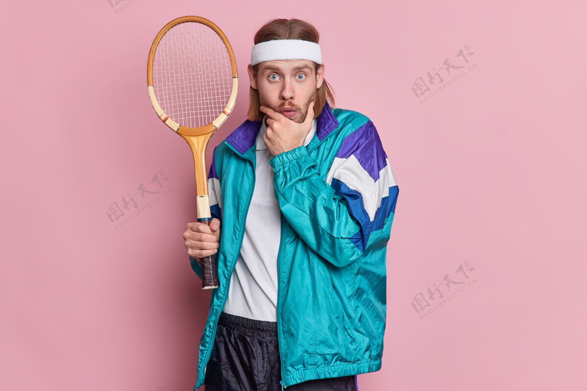 比赛惊讶的活跃的网球运动员拿着球拍看起来惊讶的握着下巴有着浓密的鬃毛戴着头带和运动服情感惊喜反应