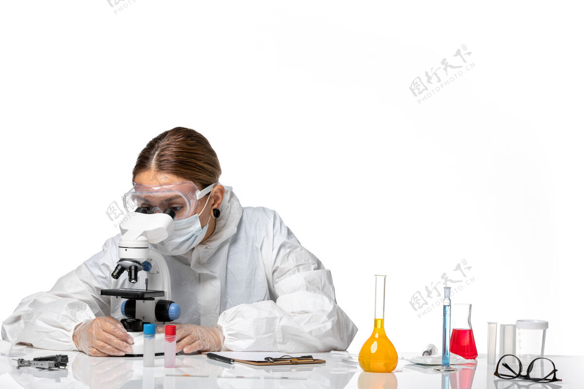 病毒前视图女医生穿着特制的西装 戴着口罩 用显微镜观察白色背景上的冠状病毒大流行实验室外套女医生面具