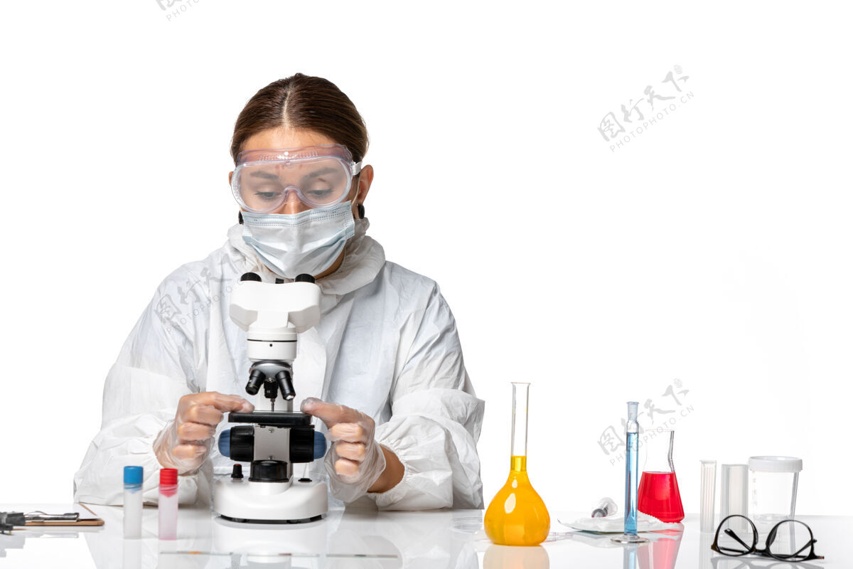 西装前视图女医生穿着特制的工作服 戴着口罩 用显微镜观察浅白色背景的冠状病毒大流行工作视图实验室